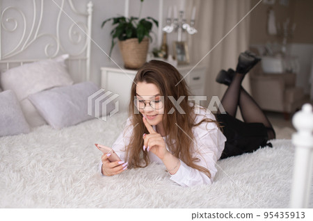 A young woman with glasses is lying on the bed and reading the news or sending a text message. 95435913