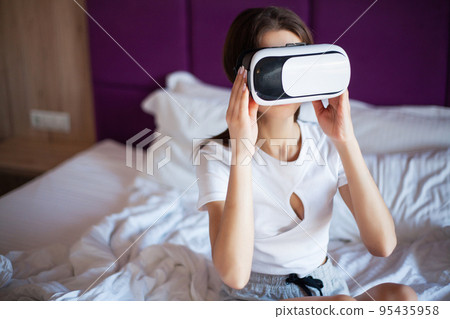 Cute woman sitting on king-size bed plays VR games in hotel room Cute woman sitting on king-size bed plays VR games in hotel room 95435958