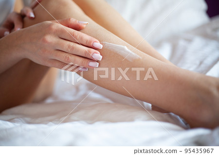 Woman applying body cream on her leg in bedroom Woman applying body cream on her leg in bedroom 95435967