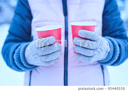 A woman in gloves holds in her hands two red paper cups of hot drink in the winter on the nature, a hot tea in the cold season, a walk and a small trip in the winter. High quality photo A woman in gloves holds in her hands two red paper cups of hot drink in the winter on the nature, a hot tea in the cold season, a walk and a small trip in the winter. High quality photo 95440550