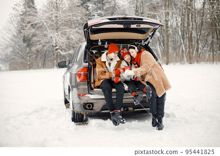 Mother, teens brother and sister spending time together in the winter park. Family sitting in open car's trunk and holding red cups with tea. Family wearing warm jackets and knitted hats. Mother, teens brother and sister spending time together in the winter park. Family sitting in open car's trunk and holding red cups with tea. Family wearing warm jackets and knitted hats. 95441825