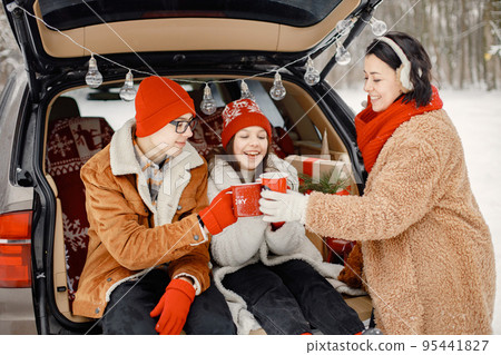 Mother, teens brother and sister spending time together in the winter park. Family sitting in open car's trunk and holding red cups with tea. Family wearing warm jackets and knitted hats. Mother, teens brother and sister spending time together in the winter park. Family sitting in open car's trunk and holding red cups with tea. Family wearing warm jackets and knitted hats. 95441827