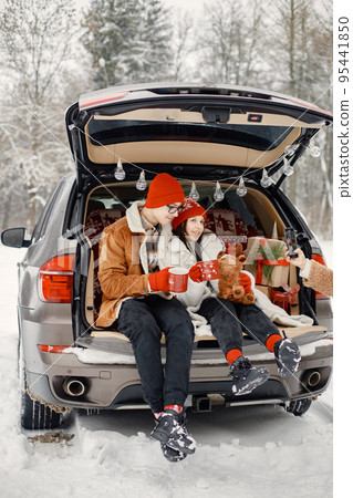 Joyful teens brother and sister spending time together in the winter park. Boy and girl sitting in open car's trunk and holding red cups with tea. Family wearing warm jackets and knitted hats. 95441850