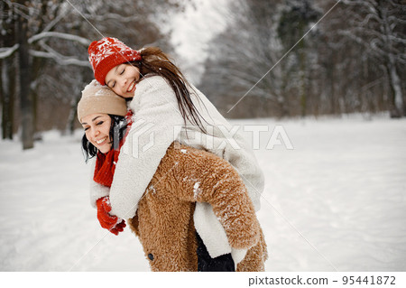 Joyful girl and her beautiful mother spending time together in the winter park. Woman wearing warm jacket and red sweater. Mother carrying her daughter piggyback. Joyful girl and her beautiful mother spending time together in the winter park. Woman wearing warm jacket and red sweater. Mother carrying her daughter piggyback. 95441872