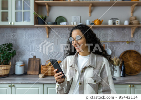 Hispanic woman uses mobile phone at home in the kitchen, woman smiles and rejoices, reads messages, browses web pages, online communication with friends and dating. 95442114
