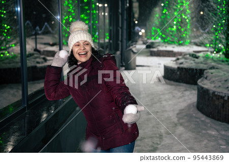 Attractive Hispanic woman wearing warm clothes, laughing, smiling, having fun in a city street, illuminated by garlands, playing with snowballs on a winter snowy evening. Christmas holidays concept Attractive Hispanic woman wearing warm clothes, laughing, smiling, having fun in a city street, illuminated by garlands, playing with snowballs on a winter snowy evening. Christmas holidays concept 95443869
