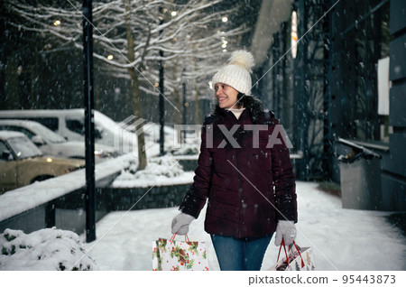 Delightful Hispanic woman in warm clothes, walking down the city street, illuminated by garlands, with shopping bags in her hands, on a snowy winter night. Merry Christmas and Happy New Year concept 95443873
