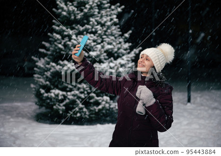 Multi-ethnic pretty woman in warm clothes, white wool mittens and hat, smiles a beautiful toothy smile while taking selfie on her smartphone, enjoying a snowy winter evening. Merry Christmas concept 95443884