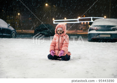 Happy Caucasian child, preschooler little girl in warm winter pink overalls sits on a snowy street, enjoying the first snow during an evening walk. Winter. Snow covered city. Childhood. 95443890