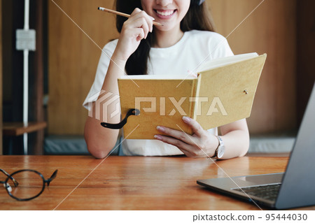 Portrait of a teenage Asian woman using a computer, wearing headphones and using a notebook to study online via video conferencing on a wooden desk in library. Portrait of a teenage Asian woman using a computer, wearing headphones and using a notebook to study online via video conferencing on a wooden desk in library. 95444030