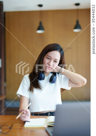 A portrait of a young Asian woman using a computer, wearing headphones and using a notebook to study online shows boredom and pain from video conferencing on a wooden desk in library. 95444032