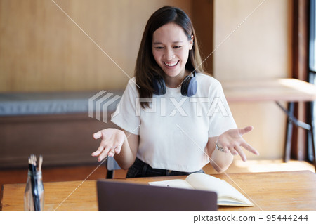 Portrait of a teenage Asian woman using a computer and notebook to study online via video conferencing on a wooden desk in library. Portrait of a teenage Asian woman using a computer and notebook to study online via video conferencing on a wooden desk in library. 95444244