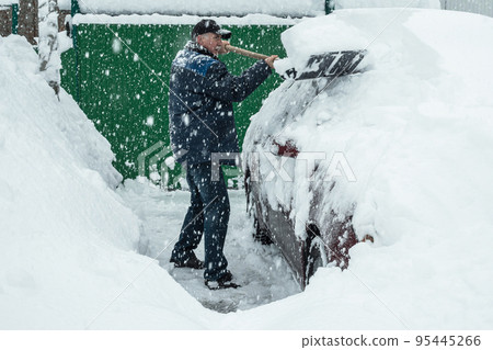 A man in a cap is shoveling snow off a car that was covered in snow during the snowfall. In the background is a green gate. Large flakes of snow are falling. A snowy winter in Europe. A man in a cap is shoveling snow off a car that was covered in snow during the snowfall. In the background is a green gate. Large flakes of snow are falling. A snowy winter in Europe. 95445266