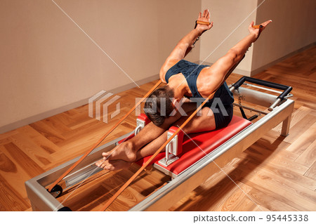 Fit man doing a lunge stretch yoga pilates exercise to strengthen and tone his muscles using a reformer in a gym Fit man doing a lunge stretch yoga pilates exercise to strengthen and tone his muscles using a reformer in a gym 95445338
