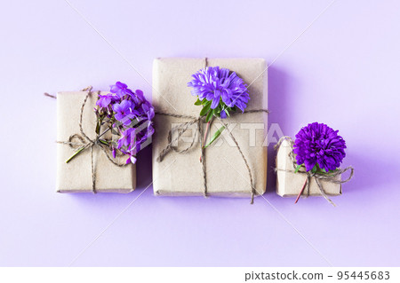 Three gift boxes in gray craft paper decorated with lilac aster flowers on a purple background for the holiday, front view Three gift boxes in gray craft paper decorated with lilac aster flowers on a purple background for the holiday, front view 95445683