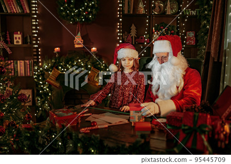 Santa Clause packs gifts with his young girl helper while sitting at the table near fireplace. Santa Clause packs gifts with his young girl helper while sitting at the table near fireplace. 95445709