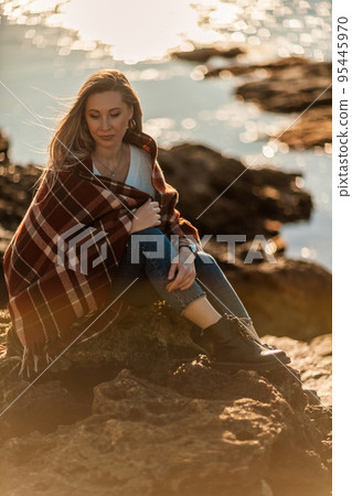 Attractive blonde Caucasian woman enjoying time on the beach at sunset, sitting in a blanket and looking to the side, with the sunset sky and sea in the background. Beach vacation. Attractive blonde Caucasian woman enjoying time on the beach at sunset, sitting in a blanket and looking to the side, with the sunset sky and sea in the background. Beach vacation. 95445970