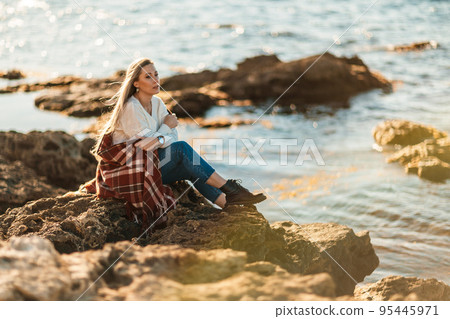 Attractive blonde Caucasian woman enjoying time on the beach at sunset, sitting in a blanket and looking to the side, with the sunset sky and sea in the background. Beach vacation. Attractive blonde Caucasian woman enjoying time on the beach at sunset, sitting in a blanket and looking to the side, with the sunset sky and sea in the background. Beach vacation. 95445971