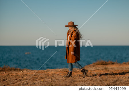 A woman walking along the coast near the sea. An elegant lady in a brown coat and a hat with fashionable makeup walks on the seashore A woman walking along the coast near the sea. An elegant lady in a brown coat and a hat with fashionable makeup walks on the seashore 95445999