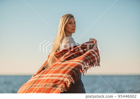 Attractive blonde Caucasian woman enjoying time on the beach at sunset, walking in a blanket and looking to the side, with the sunset sky and sea in the background. Beach vacation Attractive blonde Caucasian woman enjoying time on the beach at sunset, walking in a blanket and looking to the side, with the sunset sky and sea in the background. Beach vacation 95446002