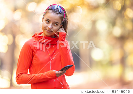 Young female athlete in sportswear listens to music on smartphone and puts headphones before running Young female athlete in sportswear listens to music on smartphone and puts headphones before running 95446046
