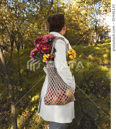 a young woman with a woven thread bag on her shoulder, dressed in a white coat, walks in the autumn park. Macrame bag with flowers and apples a young woman with a woven thread bag on her shoulder, dressed in a white coat, walks in the autumn park. Macrame bag with flowers and apples 95446145