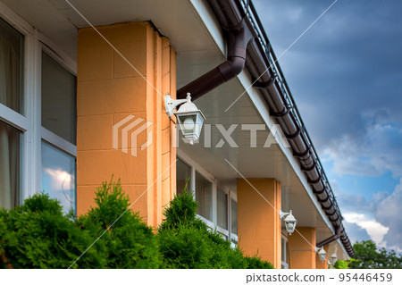 a white iron outdoor lantern with a bulb on a column of the facade of the building with a visor at the windows and a storm gutter to drain rainwater downspout from the roof, thuja bushes near house. 95446459