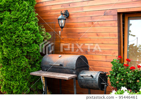 iron black rusty barbecue near wooden house with retro lantern on wall made of boards, cottage with thuja bushes and red pelargonium flowers, place to relax and cook meat and vegetables on grill. iron black rusty barbecue near wooden house with retro lantern on wall made of boards, cottage with thuja bushes and red pelargonium flowers, place to relax and cook meat and vegetables on grill. 95446461