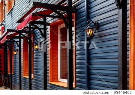 wooden facade of building with retro lanterns illuminating the house with windows and red visor, the architecture of the building in Japanese style sheathed in blue molding, evening lighting glow. wooden facade of building with retro lanterns illuminating the house with windows and red visor, the architecture of the building in Japanese style sheathed in blue molding, evening lighting glow. 95446462