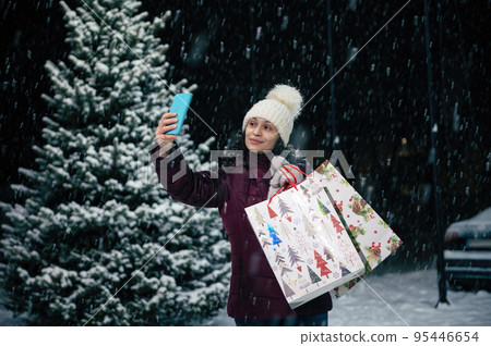 Charming woman taking a selfie on her smartphone while standing with shopping bags on the background of a snow-covered fir tree on a snowy winter evening. Merry Christmas concept 95446654