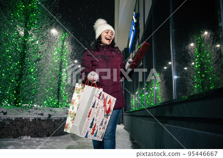 Cheerful multi-ethnic woman with a present box and shopping bags, walking down the city street illuminated by garlands, in the snowy winter evening. Christmas concept. New Year. Winter holidays Cheerful multi-ethnic woman with a present box and shopping bags, walking down the city street illuminated by garlands, in the snowy winter evening. Christmas concept. New Year. Winter holidays 95446667
