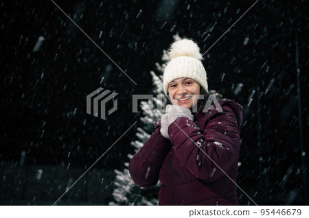 Portrait of a charming dark-haired multi-ethnic woman in warm clothes, white wool hat and mittens, smiling a beautiful toothy smile looking at the camera, standing in the snow covered nature. Portrait of a charming dark-haired multi-ethnic woman in warm clothes, white wool hat and mittens, smiling a beautiful toothy smile looking at the camera, standing in the snow covered nature. 95446679