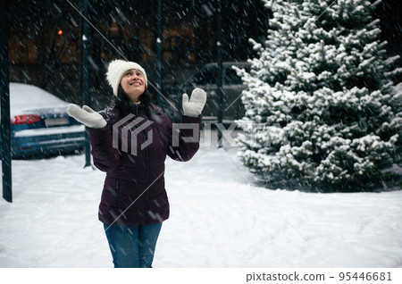 Charming cheerful multi-ethnic woman wearing wool mittens and warm clothes, looking up the evening sky, enjoys the snowfall in the city Charming cheerful multi-ethnic woman wearing wool mittens and warm clothes, looking up the evening sky, enjoys the snowfall in the city 95446681