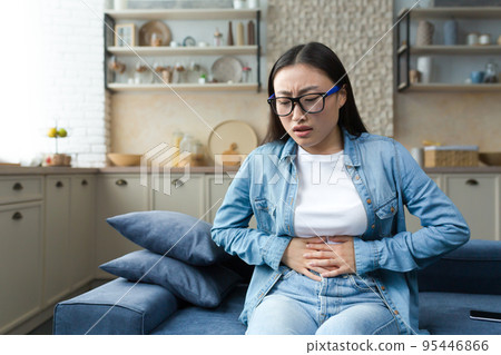 Stomach disorder. A young beautiful Asian woman holds her stomach with her hands, feels severe pain. Sitting at home on the sofa. Stomach disorder. A young beautiful Asian woman holds her stomach with her hands, feels severe pain. Sitting at home on the sofa. 95446866