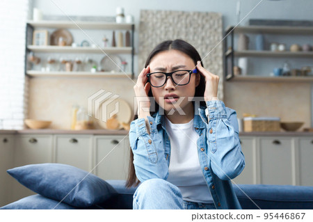 Headache, exhaustion. A young beautiful Asian woman holds her head, feels pain, pressure, tension. Sitting on the sofa, at home. Needs help. Headache, exhaustion. A young beautiful Asian woman holds her head, feels pain, pressure, tension. Sitting on the sofa, at home. Needs help. 95446867