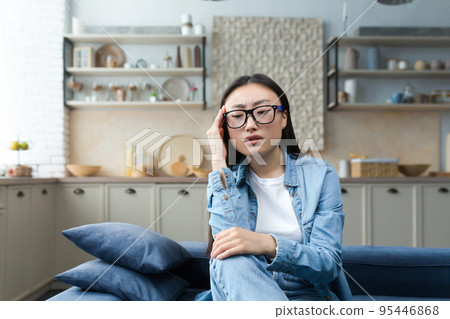 Depression. Tired and pensive young Asian woman sitting on sofa at home, holding hand to head, looking focused. Depression. Tired and pensive young Asian woman sitting on sofa at home, holding hand to head, looking focused. 95446868