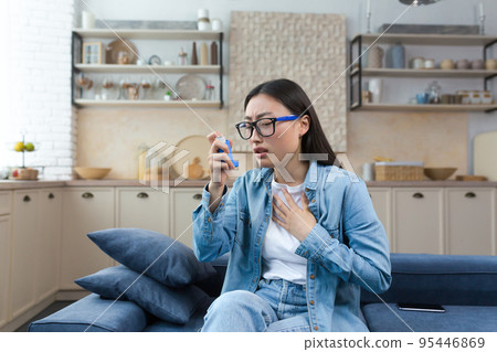 Allergic attack. Asthma. Young Asian woman sitting on sofa at home and using inhaler. Has an asthma attack, treats allergies, has difficulty breathing. Allergic attack. Asthma. Young Asian woman sitting on sofa at home and using inhaler. Has an asthma attack, treats allergies, has difficulty breathing. 95446869
