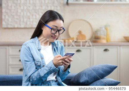 A happy young beautiful Asian woman is sitting on the sofa at home. She is holding a mobile phone in her hands, writing a message. Corresponds with boyfriend, friends, smiles A happy young beautiful Asian woman is sitting on the sofa at home. She is holding a mobile phone in her hands, writing a message. Corresponds with boyfriend, friends, smiles 95446870
