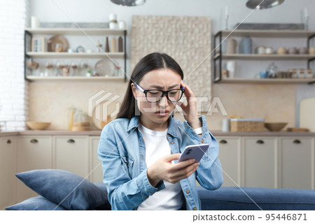 Worried young Asian woman holds the phone in her hands, received bad news by message. Sitting at home on the sofa, holding her head, nervous. Worried young Asian woman holds the phone in her hands, received bad news by message. Sitting at home on the sofa, holding her head, nervous. 95446871