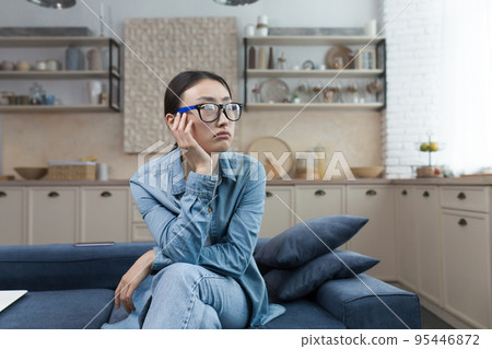 Depression. Pensive young beautiful Asian woman sitting on the sofa at home, looking out the window, resting her head on her hand, having a bad mood, anxious thoughts. Depression. Pensive young beautiful Asian woman sitting on the sofa at home, looking out the window, resting her head on her hand, having a bad mood, anxious thoughts. 95446872