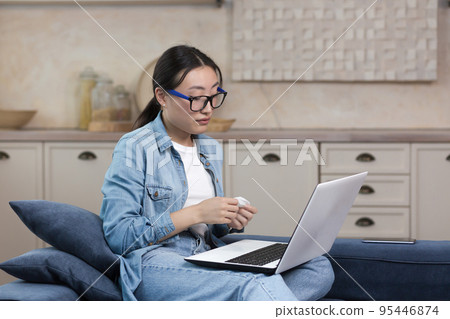 A young beautiful Asian woman is sitting at home on the couch, watching a movie on a laptop, crying, holding a napkin in her hands. Touched, spends the evening alone. A young beautiful Asian woman is sitting at home on the couch, watching a movie on a laptop, crying, holding a napkin in her hands. Touched, spends the evening alone. 95446874