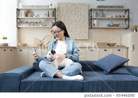 A happy young beautiful Asian woman in glasses is talking on a video call, holding a phone and her dog in her hands, smiling at the camera. Sitting at home on the sofa in the lotus position. A happy young beautiful Asian woman in glasses is talking on a video call, holding a phone and her dog in her hands, smiling at the camera. Sitting at home on the sofa in the lotus position. 95446890
