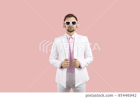 Fashionable confident man in a white classic suit and a pink shirt in sunglasses with headphones on a pink background. 95446942