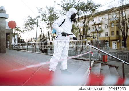 Man in protective suit and mask sprays disinfector onto the railing in the public place. Covid -19. Man in protective suit and mask sprays disinfector onto the railing in the public place. Covid -19. 95447063