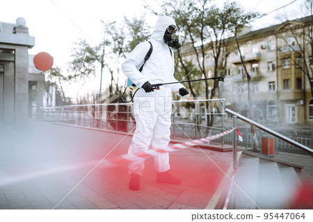 Man in protective suit and mask sprays disinfector onto the railing in the public place. Covid -19. 95447064