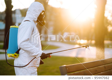 Man wearing protective suit disinfecting public places in the sun with spray chemicals. Covid -19. 95447082
