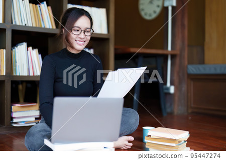 A portrait of a young Asian female student with a smiling face using a computer and holding study materials in the library. A portrait of a young Asian female student with a smiling face using a computer and holding study materials in the library. 95447274