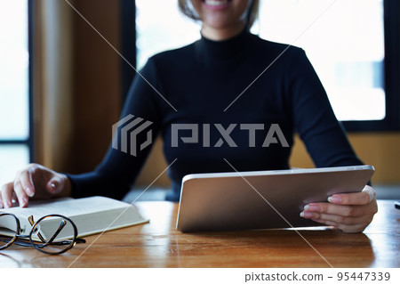 Portrait of a teenage Asian woman using a tablet computer and books to study online via video conferencing on a wooden table in the library. Portrait of a teenage Asian woman using a tablet computer and books to study online via video conferencing on a wooden table in the library. 95447339