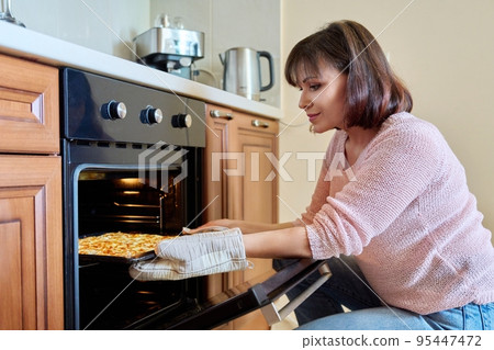 Middle-aged woman with cooked meat taking baking sheet out of oven 95447472