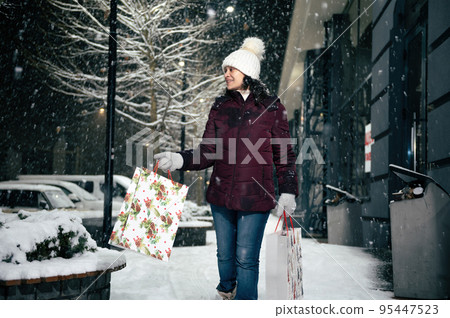 Delightful happy young multi-ethnic woman walking down the street with shopping bags, on a snowy winter cool evening. Merry Christmas. 95447523